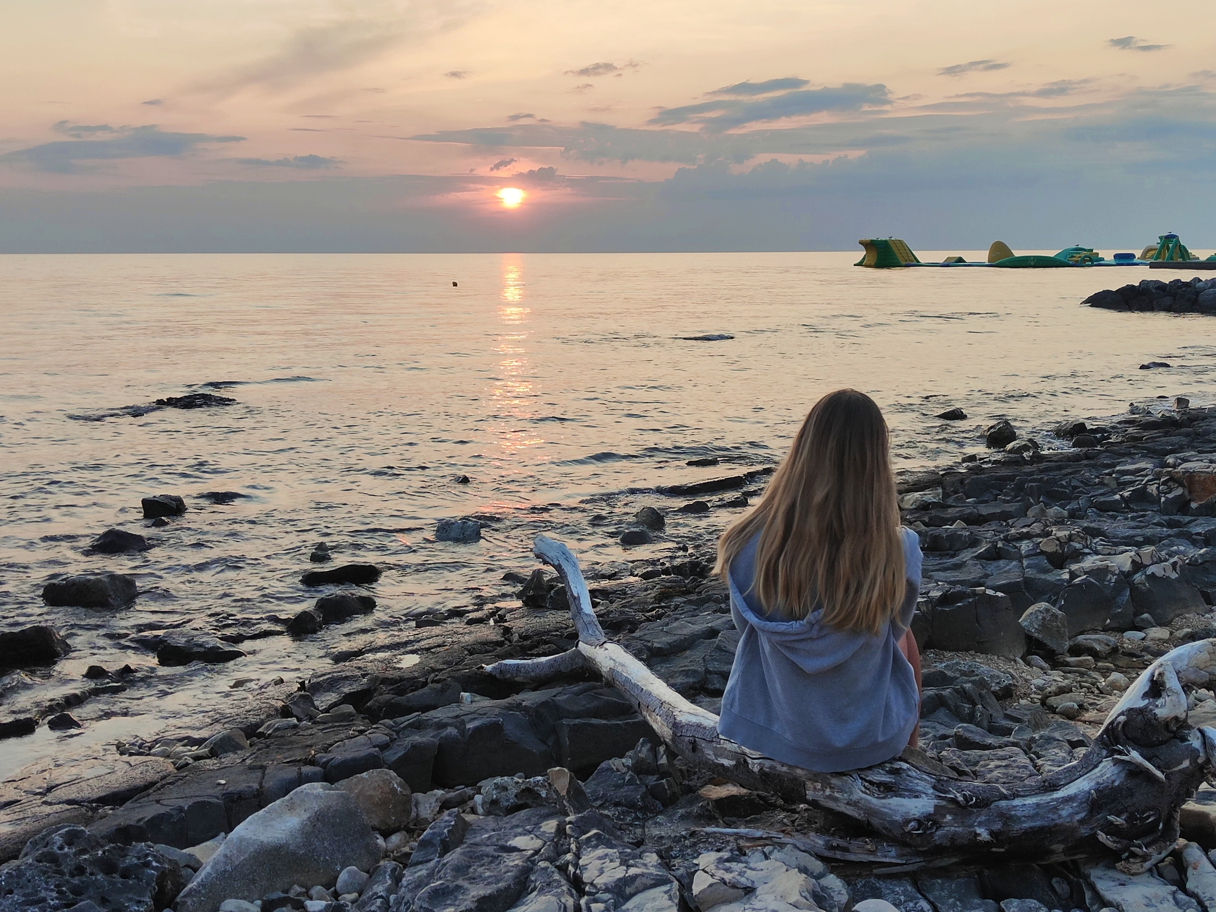 Man sieht einen Steinstrand in Kroatien im Sonnenuntergang. Auf einem Felsen am Meer sieht man ein Mädchen mit langen Haaren von hinten, das übers Meer schaut.
