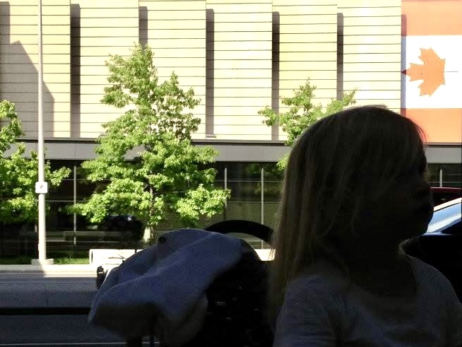 Im Vordergrund ein Kind. das auf einem Koffer sitzt und die USA über Kanada bereist. Im Hintergrund ein Gebäude mit kanadischer Flagge.